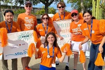 strom thacker poses with student admission staff on move in day 2023. Everyone is wearing orange pitzer t-shirts and holding pom poms.